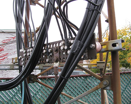 Figure 5. A copper grounding busbar on the tower (center of photo) connects grounded shields on waveguides and coax cable shields serving various antennas (smaller-diameter cables). The large-diameter, vertical green grounding cable visible at the far lower left corner connects the busbar with the three driven electrodes and the buried copper ring ground surrounding the tower.