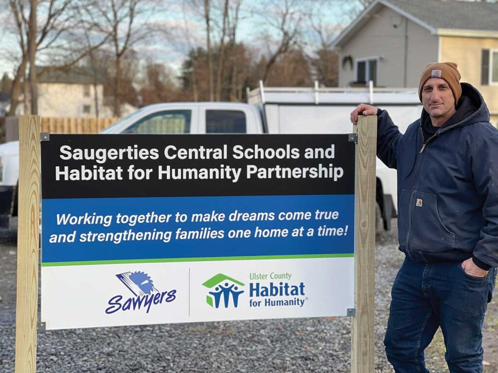 Photo 2. Saugerties Guidance Counselor and lead project advisor Michael Catalano stands out in front of a sign by the three homes Ulster BOCES students have volunteered to help build. Photo Courtesy-Kristine Conte/Ulster BOCES